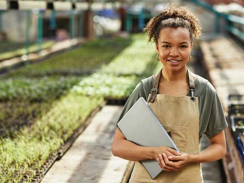 Confident young grower standing in front of her seedlings, holding her laptop.