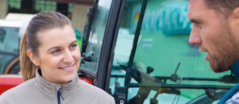 A woman standing smiling and holding clipboard as a male farmer talks to her.