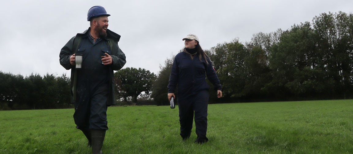 Farmer walks grass field with new employee, joking about the poor weather.